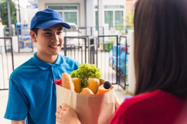 Üniformalı Asyalı genç teslimatçı market hızlı servisini yapıyor. Kadın müşterilere, pandemik koronavirüs altında evlerinin kapısında taze yiyecek veriyor. Yeni normal konsepte dönüyoruz.