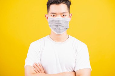 Yüzüne koruyucu mikrop takan yakışıklı Asyalı genç adam çapraz kol duruşu, sarı arka plan ve fotokopi alanında izole edilmiş stüdyo çekimi, tıbbi salgın Coronavirus COVID-19 konsepti.