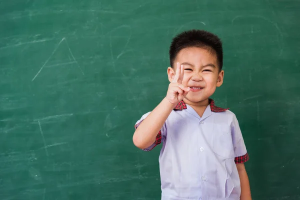 Back to School. Happy Asian funny cute little child boy from kindergarten in student uniform show finger V-sign smiling on green school blackboard, First time to school education concept, studio shot