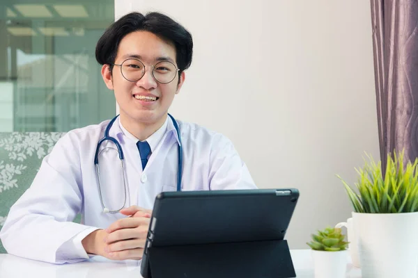 幸せなアジアの若い医師ハンサムな男の笑顔眼鏡を使用して現代的なスマートデジタルタブレットコンピュータ座って 患者が病院のオフィスで机の上に症状を説明聞きます テクノロジーヘルスケアと医学 — ストック写真