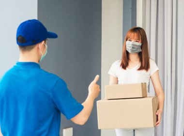 Asyalı kurye genç adam kadın müşteriye paket kutuları veriyor. Hem koruyucu yüz maskesi alıyor hem de sokağa çıkma yasağı pandemik koronavirüs altında iyi bir destek işareti için parmaklarını gösteriyor.