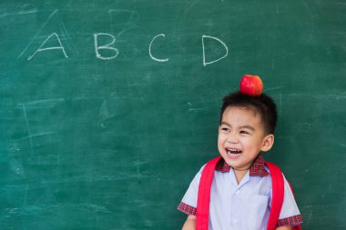Okula geri dön. Anaokulundan, okul çantasıyla, kafasında kırmızı elmayla yeşil okul tahtasına gülümseyen küçük, mutlu Asyalı çocuk.