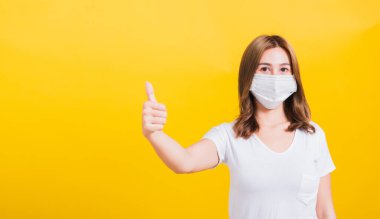 Asya Tayland portresi, maske takan güzel, mutlu genç kadın, başparmağını göster virüs salgınını önleme sağlık hizmetleri, sarı arka planda izole edilmiş stüdyo çekimi, fotokopi alanı