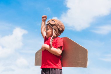 Mutlu Asyalı komik çocuk ya da küçük çocuk gülümser pilot şapkası takar ve gözlük takar oyuncak karton uçak kanatları uçar yaz mavi gökyüzü bulutu arka planına el kaldırır, başlangıç özgürlüğü kavramı