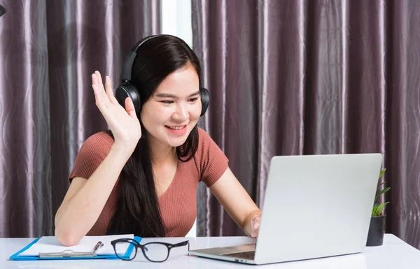 家庭からの仕事 笑顔の幸せなアジアのビジネス若い美しい女性が机の上に座っているヘッドフォンのビデオ通話会議を着てラップトップコンピュータと手を上げるホームオフィスでハイチームと言って — ストック写真