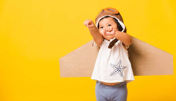 幸せなアジアの美しい面白い赤ちゃんの女の子の笑顔は おもちゃの段ボール飛行機の翼が飛ぶと手を再生し ゴーグルを上げるパイロットの帽子を身に着けている スタジオショット孤立黄色の背景 スタートアップの自由コンセプト — ストック写真