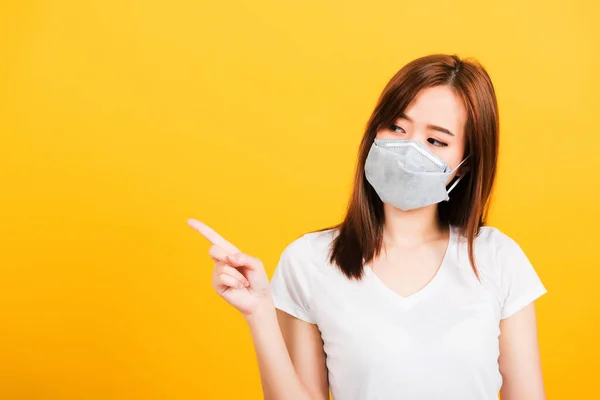 Portrait Asian beautiful happy young woman wearing face mask protects filter dust pm2.5 anti-pollution, anti-smog, and air pollution her pointing out to side on yellow background, with copy space