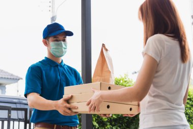Asyalı kurye genç adam, sokağa çıkma yasağı altındaki karantina altındaki kadın müşterilere fast food ve pizza kutusu veriyor.