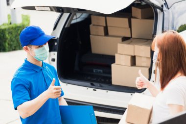 Asyalı kurye genç adam kadın müşteriye paket kutuları veriyor. Hem koruyucu yüz maskesi alıyor hem de sokağa çıkma yasağı pandemik koronavirüs altında iyi bir destek işareti için parmaklarını gösteriyor.