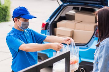 Asyalı genç teslimatçı yüz maskesi takıyor. Bakkal servisi yapıyor. Kadın müşterilere plastik torbalar veriyor.