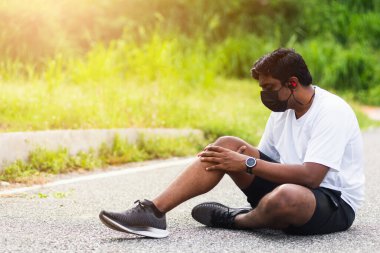 Asyalı spor koşucusu siyah adam kol saati takıyor. Açık hava parkında koşarken dizine eklem yapıyor. Egzersiz konseptinden kaynaklanan sağlıklı egzersiz sakatlığı.