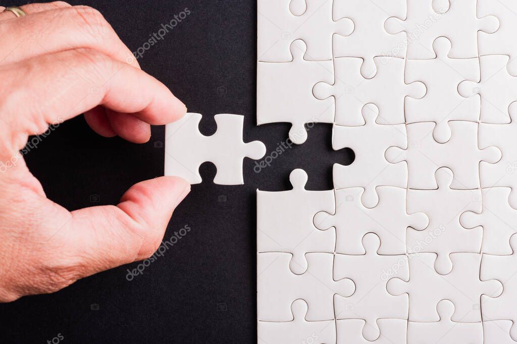 Top view flat lay of hand-holding last piece white paper jigsaw puzzle game last pieces put to place for solve problem complete mission, studio shot on a black background, quiz calculation concept