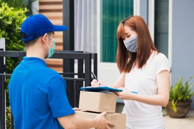 Paket postası taşıyan Asyalı genç teslimatçı, sokağa çıkma yasağı altındaki karantina altındaki kağıt defter üzerindeki kalem imzasını kullanarak koruyucu yüz maskeli kadın müşteri COVID-19 'u karantina altına aldı.