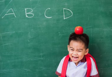 Okula geri dön. Anaokulundan, okul çantasıyla, kafasında kırmızı elmayla yeşil okul tahtasına gülümseyen küçük, mutlu Asyalı çocuk.
