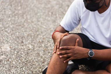 Asyalı spor koşucusu siyah adam kol saati takıyor. Açık hava parkında koşarken dizine eklem yapıyor. Egzersiz konseptinden kaynaklanan sağlıklı egzersiz sakatlığı.