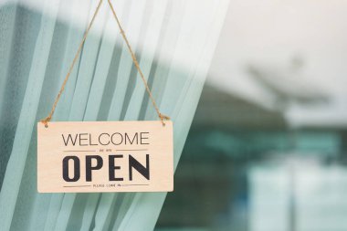 Close up of WELCOME WE ARE OPEN PLEASE COME IN notice sign wood board label hanging through glass door front shop, Business open back to new normal after coronavirus disease concept clipart