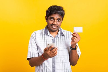 Yakın plan Asyalı mutlu portre genç siyah adam cep telefonu tutuyor ve internet üzerinden mobil ödeme konsepti için kredi kartı gösteriyor, sarı arka planda izole edilmiş stüdyo.