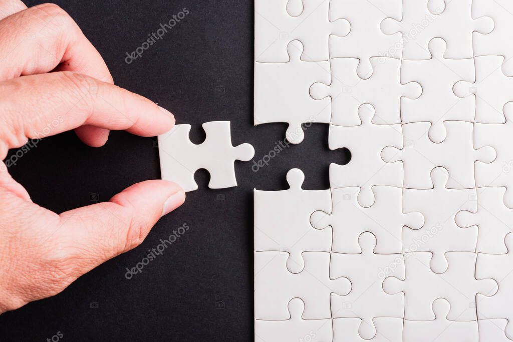 Top view flat lay of hand-holding last piece white paper jigsaw puzzle game last pieces put to place for solve problem complete mission, studio shot on a black background, quiz calculation concept