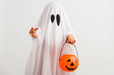 Komik Cadılar Bayramı Çocuğu kavramı, beyaz kostümlü küçük şirin çocuk Cadılar Bayramı hayaleti korkunç elinde turuncu kabak hayaleti, beyaz arka planda tek başına çekilmiş bir stüdyo resmi.