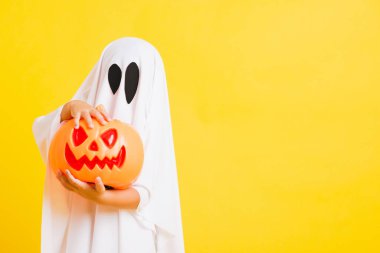 Komik cadılar bayramı çocuğu kavramı, beyaz kostümlü şirin çocuk Cadılar Bayramı hayaleti korkunç elinde turuncu kabak hayaleti, beyaz arka planda sarı bir stüdyo.