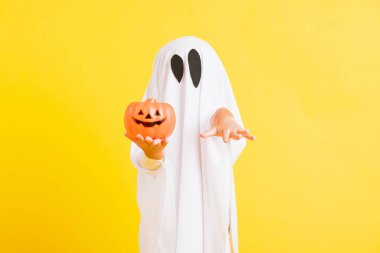 Komik cadılar bayramı çocuğu kavramı, beyaz kostümlü şirin çocuk Cadılar Bayramı hayaleti korkunç elinde turuncu kabak hayaleti, beyaz arka planda sarı bir stüdyo.