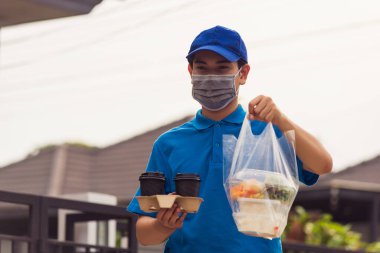 Mavi üniformalı Asyalı genç teslimatçı market servisi yapıyor. Salgın koronavirüs altında evdeki pirinç kutularına plastik torbalar ve kahve veriyor. Yeni normal konsepte geri dönüyoruz.