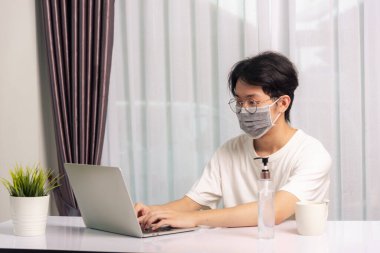 Asyalı iş adamı yakışıklı erkek gözlükleri takıyor. Yanında dezenfektan jeli koruyucu maske takıyor. Ev bilgisayarının uzaktan kumandasıyla bilgisayar karantinası Coronavirus veya COVID-19 ile çalışıyor.