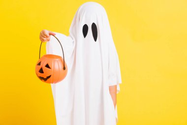 Komik cadılar bayramı çocuğu kavramı, beyaz kostümlü şirin çocuk Cadılar Bayramı hayaleti korkunç elinde turuncu kabak hayaleti, beyaz arka planda sarı bir stüdyo.