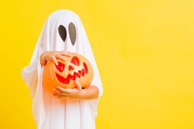 Komik cadılar bayramı çocuğu kavramı, beyaz kostümlü şirin çocuk Cadılar Bayramı hayaleti korkunç elinde turuncu kabak hayaleti, beyaz arka planda sarı bir stüdyo.
