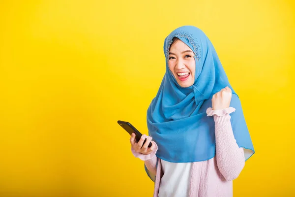 アジア系ムスリムアラブ人 幸せな美しい若い女性の肖像イスラム教の宗教的な摩耗ベールヒジャーブ面白い笑顔彼女は黄色の背景に隔離されたスマート携帯電話で受賞を祝う手を上げる — ストック写真