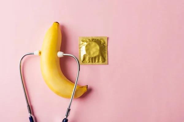 World sexual health or Aids day, Top view flat lay condom in wrapper pack, banana and doctor stethoscope, studio shot isolated on a pink background, Safe sex and reproductive health concept