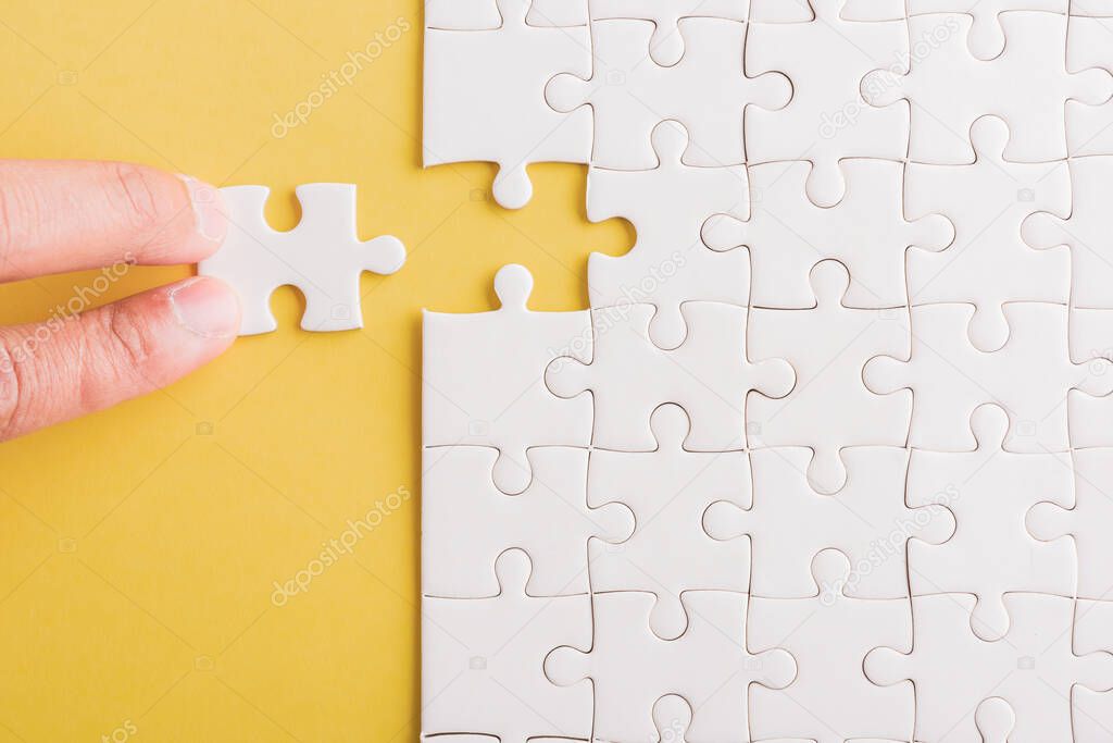 Top view flat lay of hand-holding last piece white paper jigsaw puzzle game last pieces put to place for solve problem complete mission, studio shot on a yellow background, quiz calculation concept