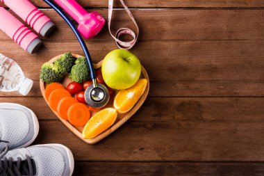 Tahta tabakta taze meyve ve sebzelerin üst görüntüsü (elma, havuç, domates, portakal, brokoli) ve spor malzemeleri ve ahşap masa üzerinde doktor steteskobu, sağlıklı yaşam tarzı diyet gıda konsepti