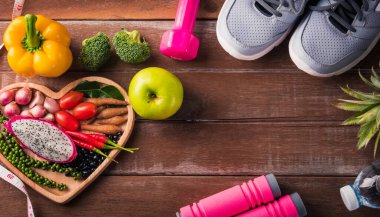 Kalp tabağında taze organik meyve ve sebze, spor ayakkabıları, dambıl ve su, ahşap spor masasında stüdyo çekimi, sağlıklı diyet vejetaryen gıda konsepti, Dünya Gıda Günü