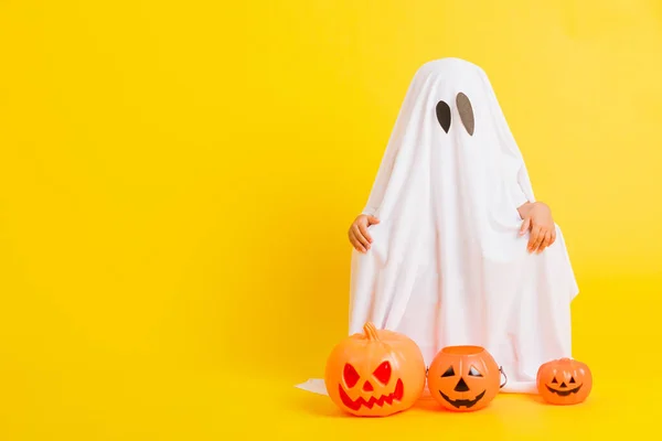 Funny Halloween Kid Concept, full body of a little cute child with white dressed costume halloween ghost scary he holding orange pumpkin ghost, studio shot yellow on white background