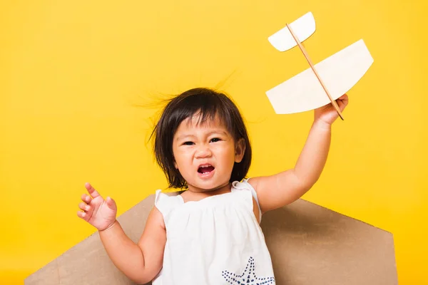幸せなアジアの美しい面白い赤ちゃんの女の子の笑顔はおもちゃの段ボール飛行機の翼でパイロットの帽子遊びを着用フライホールド飛行機のおもちゃ スタジオショット孤立した黄色の背景 スタートアップの自由の概念 — ストック写真