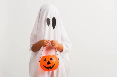 Komik Cadılar Bayramı Çocuğu kavramı, beyaz kostümlü küçük şirin çocuk Cadılar Bayramı hayaleti korkunç elinde turuncu kabak hayaleti, beyaz arka planda tek başına çekilmiş bir stüdyo resmi.