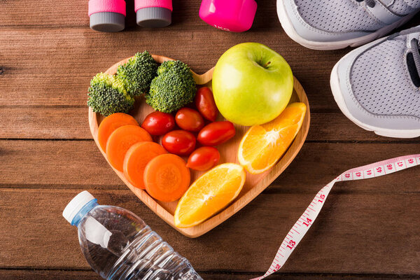 Top view of fresh fruits and vegetables in heart plate wood (apple, carrot, tomato, orange, broccoli) and dumbcoli, sport shoes sports equipment on wooden table, Healthy lifestyle diet food concept