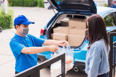 Asyalı genç teslimatçı yüz maskesi takıyor. Bakkal servisi yapıyor. Kadın müşterilere plastik torbalar veriyor.