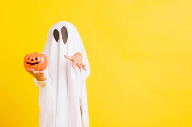 Komik cadılar bayramı çocuğu kavramı, beyaz kostümlü şirin çocuk Cadılar Bayramı hayaleti korkunç elinde turuncu kabak hayaleti, beyaz arka planda sarı bir stüdyo.