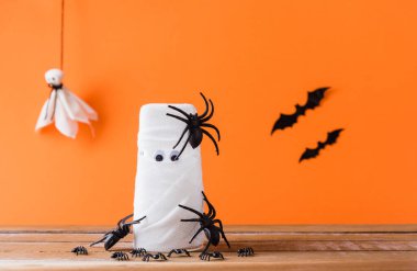 Cadılar Bayramı için komik bir dekor partisi konsepti, su bardağına sarılı mumya hayaleti, yarasalar ve örümcek sopası, sadece gözler, turuncu arka planda izole edilmiş bir stüdyo görüntüsü buldu.