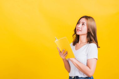 Asyalı güzel, mutlu, genç bir kadının portresi sarı kitap ya da günlüğü tutuyor başparmağını kaldırıyor ve kafasını çeviriyor, sarı arka planda izole edilmiş bir stüdyo resmi, kopyalama alanı ile birlikte.