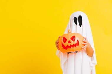 Komik cadılar bayramı çocuğu kavramı, beyaz kostümlü şirin çocuk Cadılar Bayramı hayaleti korkunç elinde turuncu kabak hayaleti, beyaz arka planda sarı bir stüdyo.
