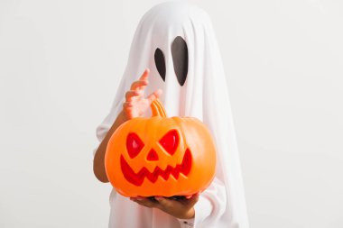 Komik Cadılar Bayramı Çocuğu kavramı, beyaz kostümlü küçük şirin çocuk Cadılar Bayramı hayaleti korkunç elinde turuncu kabak hayaleti, beyaz arka planda tek başına çekilmiş bir stüdyo resmi.