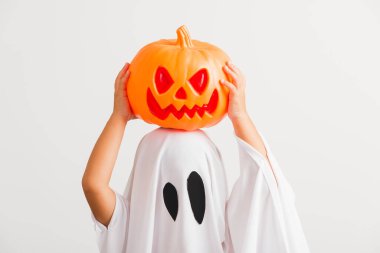 Komik Cadılar Bayramı Çocuğu kavramı, beyaz kostümlü küçük şirin çocuk Cadılar Bayramı hayaleti korkunç elinde turuncu kabak hayaleti, beyaz arka planda tek başına çekilmiş bir stüdyo resmi.