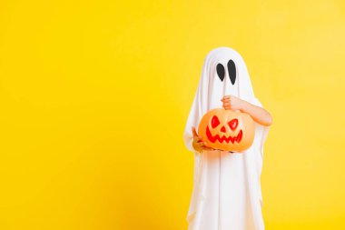 Komik cadılar bayramı çocuğu kavramı, beyaz kostümlü şirin çocuk Cadılar Bayramı hayaleti korkunç elinde turuncu kabak hayaleti, beyaz arka planda sarı bir stüdyo.