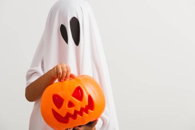 Komik Cadılar Bayramı Çocuğu kavramı, beyaz kostümlü küçük şirin çocuk Cadılar Bayramı hayaleti korkunç elinde turuncu kabak hayaleti, beyaz arka planda tek başına çekilmiş bir stüdyo resmi.