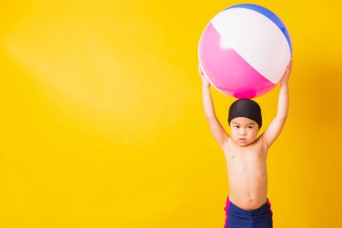Yaz tatili konsepti, Asyalı mutlu çocuk portresi, mayo içinde gülümseyen çocuk plaj topu, yaz tatilinde şişme topla eğlenen çocuk, izole edilmiş sarı arka plan.