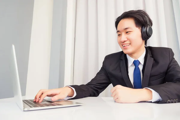 自宅からの仕事 ヘッドフォンを身に着けているアジアの若いビジネスマンの笑顔とスーツビデオ会議コールや自宅のオフィスでデスク上のラップトップコンピュータとFacetime — ストック写真