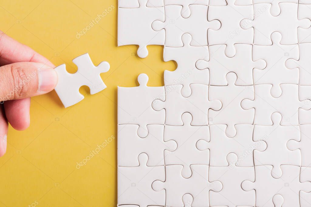 Top view flat lay of hand-holding last piece white paper jigsaw puzzle game last pieces put to place for solve problem complete mission, studio shot on a yellow background, quiz calculation concept